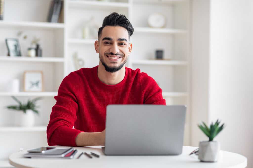 Membre LEO souriant, portant un chandail rouge, assis à un bureau avec un ordinateur portable dans un bureau à domicile lumineux et moderne.