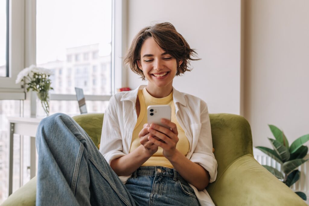 A young woman with short brown hair is sitting comfortably in a green armchair, smiling as she looks at her smartphone, reading everything she needs to know about Leo. She is wearing a yellow top under an open white shirt and blue jeans. Behind her, large windows reveal tall apartment buildings and natural light fills the room. A vase of white flowers and a potted plant are visible in the background, creating a cozy, modern atmosphere.