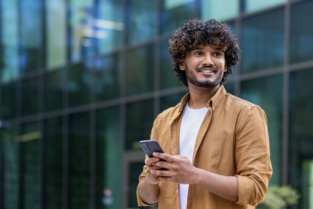 Jeune homme souriant tenant son téléphone, debout devant un immeuble moderne, consultant probablement un sondage LEO sur les enjeux électoraux de 2025.