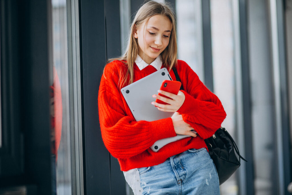 Young woman in a red sweater holding a laptop and reading the LEO blog on her smartphone at school
