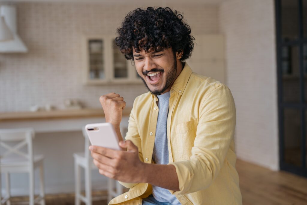 A man with curly hair in a yellow shirt cheers and pumps his fist while staring at his phone—he just placed a bet on a LEO contest and is bursting with excitement.