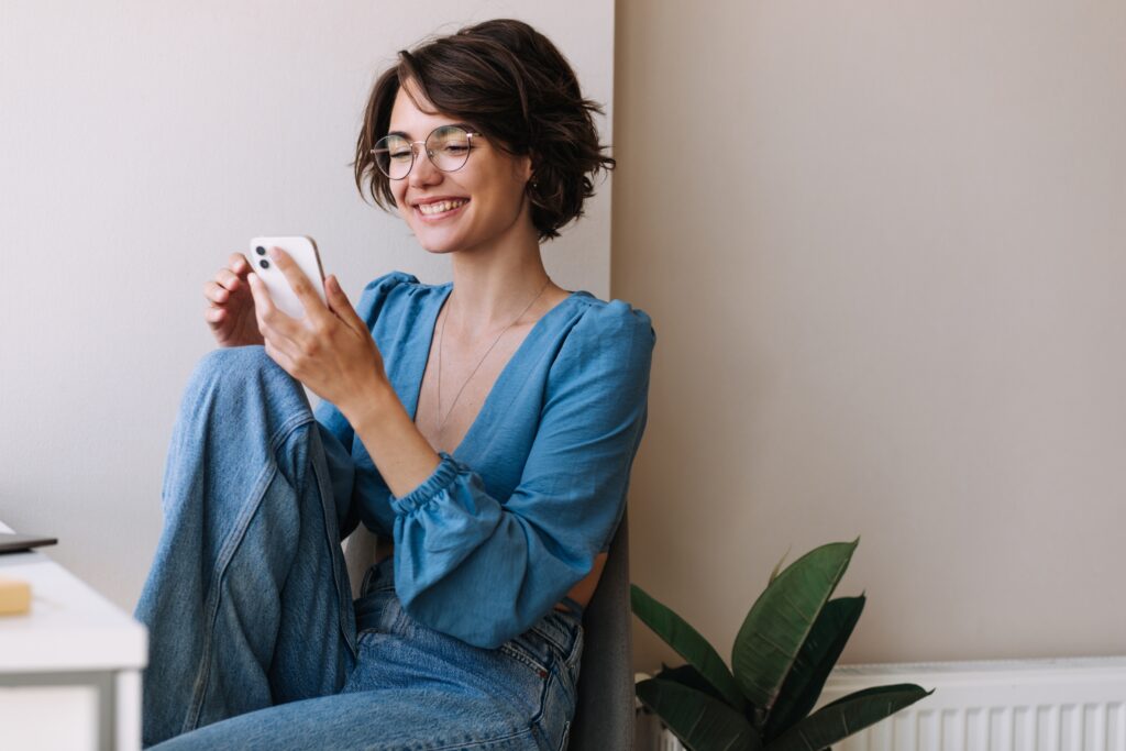 Une femme souriante, portant un chemisier bleu et des lunettes, se détend près du mur tout en regardant son téléphone — elle parcourt les derniers concours LEO avec intérêt et enthousiasme.