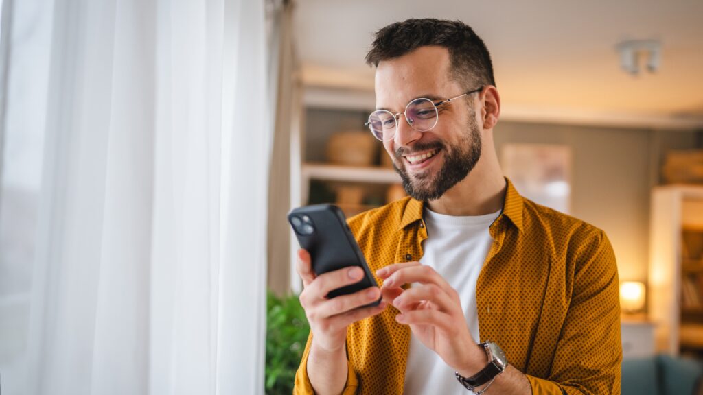 Jeune homme souriant avec des lunettes consultant les concours LEO sur son smartphone près d'une fenêtre dans un intérieur moderne et lumineux