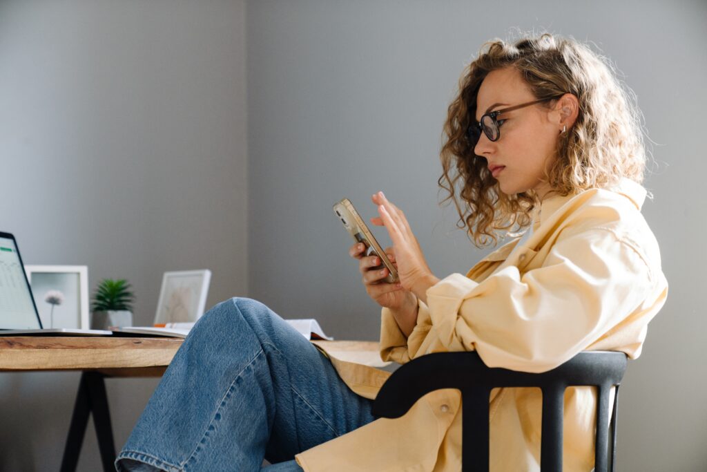 Jeune professionnelle assise à un bureau, concentrée sur son smartphone en télétravail, faisant une pause et lisant le blog Leo sur les conflits, les tarifs et les tensions.
