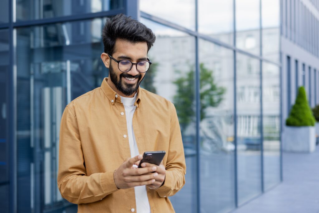 A smiling man in a mustard shirt standing outside a modern office building, looking at his smartphone — representing a user submitting questions for LEO to answer via mobile.