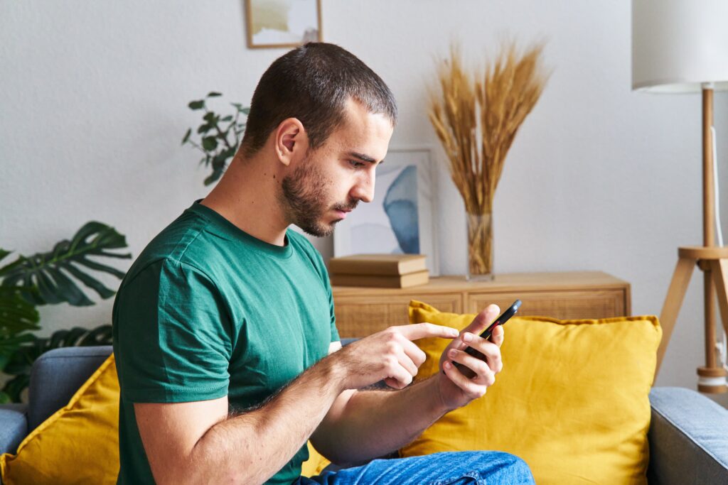Homme en t-shirt vert assis sur un canapé, concentré, remplissant des sondages sur l’application LEO à l’aide d’un téléphone intelligent à l’intérieur