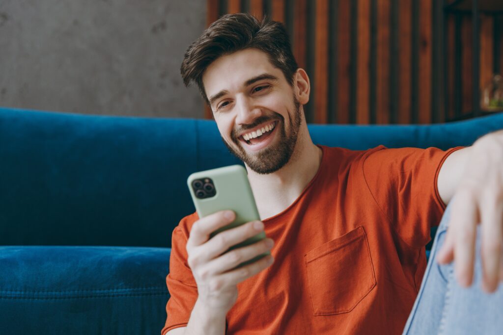 Young man smiling while sitting on a couch, using his phone to calculate his happiness index.