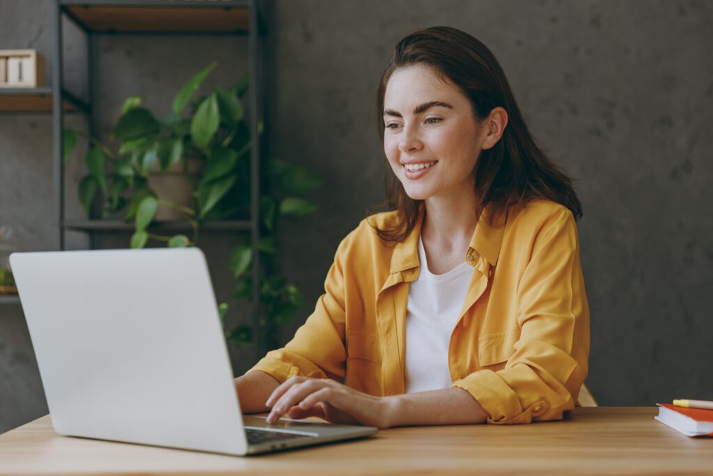 Young woman in a yellow shirt completing LEO surveys on her laptop at a desk with plants in the background.