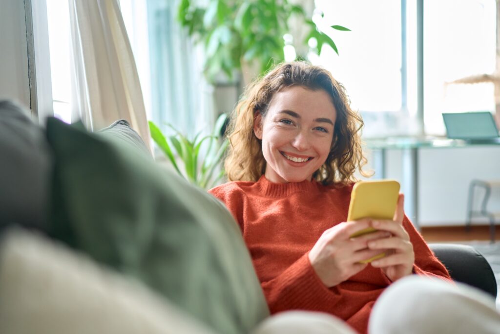 Smiling young woman relaxing on a couch while using the LEO platform on her smartphone, enjoying a seamless digital experience at home.