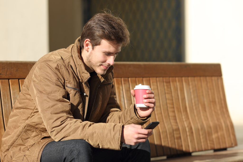 Young man sitting on a bench outdoors holding a coffee cup and using his smartphone to view LEO's October 2025 contests.