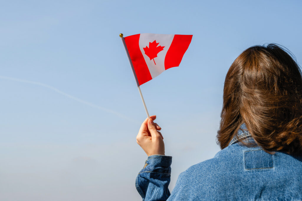 Person holding a small Canadian flag outdoors on a clear day, celebrating Canada with blue sky background.
