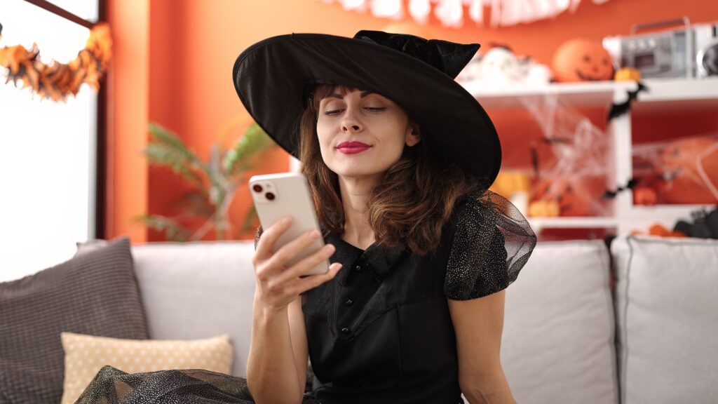 Femme souriante déguisée en sorcière utilisant son smartphone à la maison, répondant à des sondages LEO avec des décorations d’Halloween en arrière-plan.