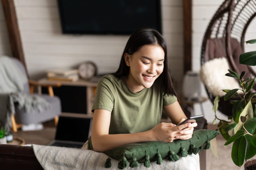 Smiling young woman sitting comfortably at home, using the LEO platform on her smartphone for an enjoyable and easy digital experience.