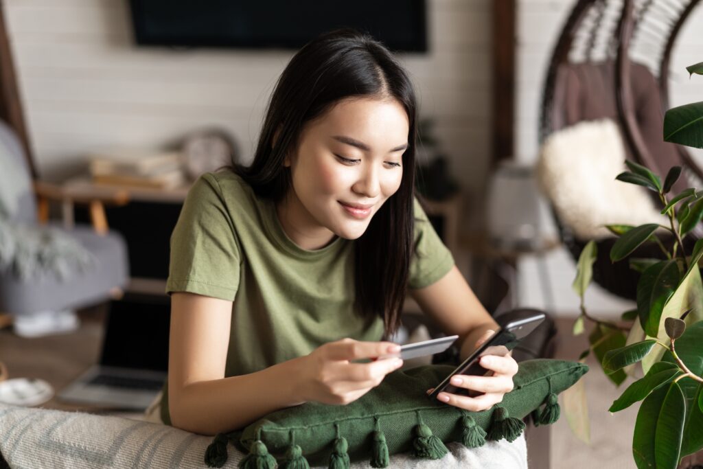 Jeune femme confortablement installée à la maison, tenant une carte-cadeau et utilisant la plateforme LEO sur son smartphone pour magasiner et échanger ses récompenses lors du Cyber Monday.