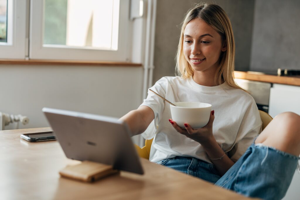 Jeune femme souriante assise à la maison avec un bol de céréales, utilisant une tablette pour répondre à un sondage LEO en ligne.