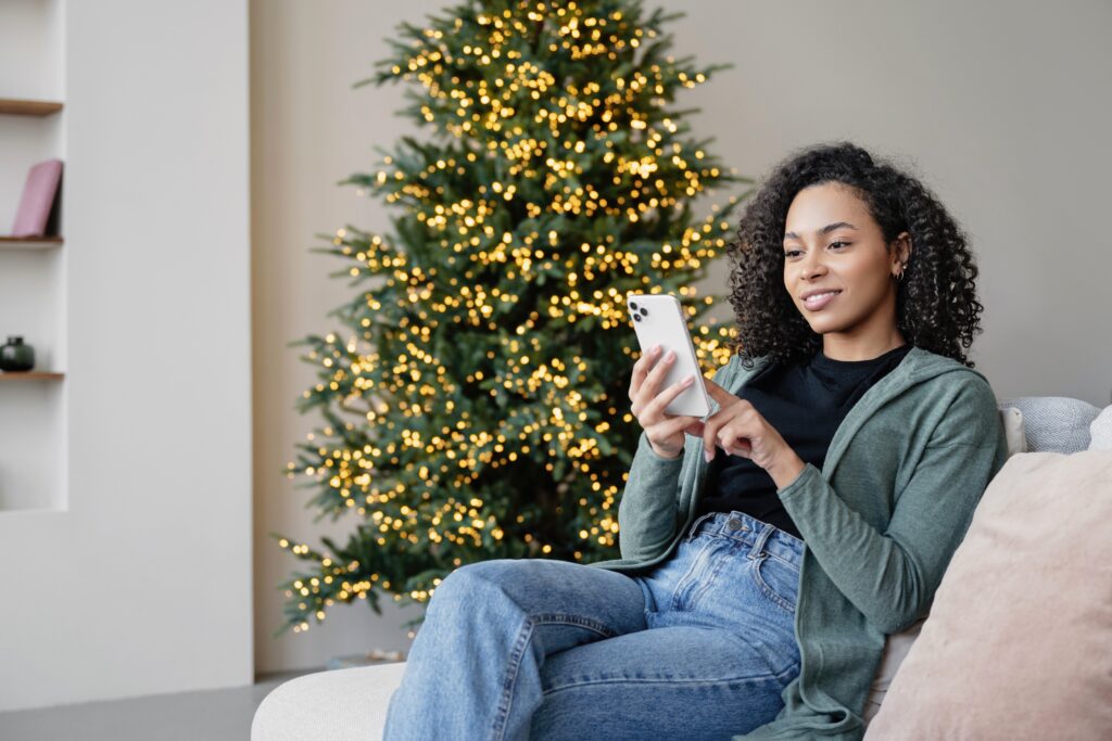 Young woman relaxing at home near a decorated Christmas tree, using the LEO platform on her smartphone for a smooth and engaging digital experience.