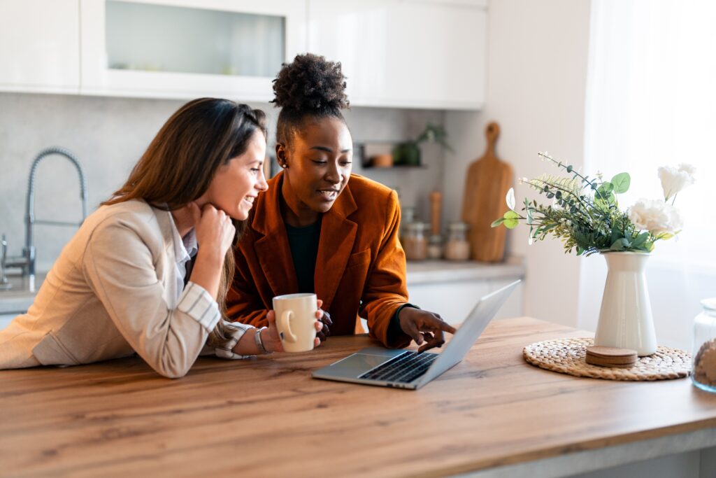 Deux femmes dans une cuisine lumineuse regardant un ordinateur portable ensemble, explorant la plateforme LEO et discutant de leur expérience numérique.