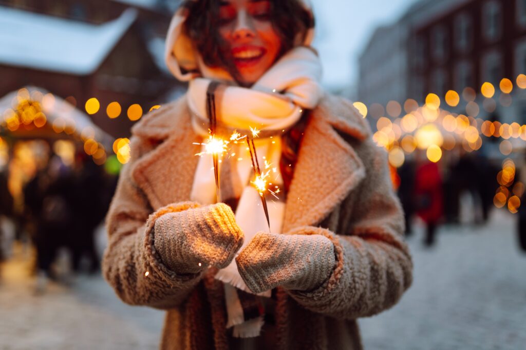 Smiling woman outdoors during a festive winter evening, holding sparklers while celebrating the end of 2025 and welcoming the beginning of 2026