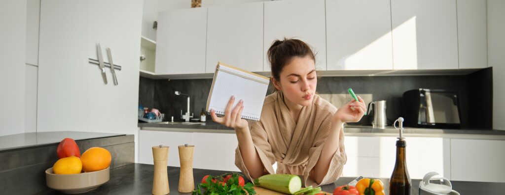 Young woman in her kitchen reviewing notes while preparing fresh ingredients, thoughtfully planning her day and incorporating the LEO platform into her daily routine.
