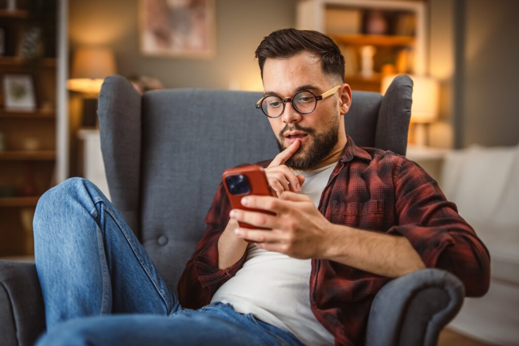 Thoughtful young man sitting in an armchair at home, using the LEO platform on his smartphone to browse surveys and rewards in a relaxed evening setting.