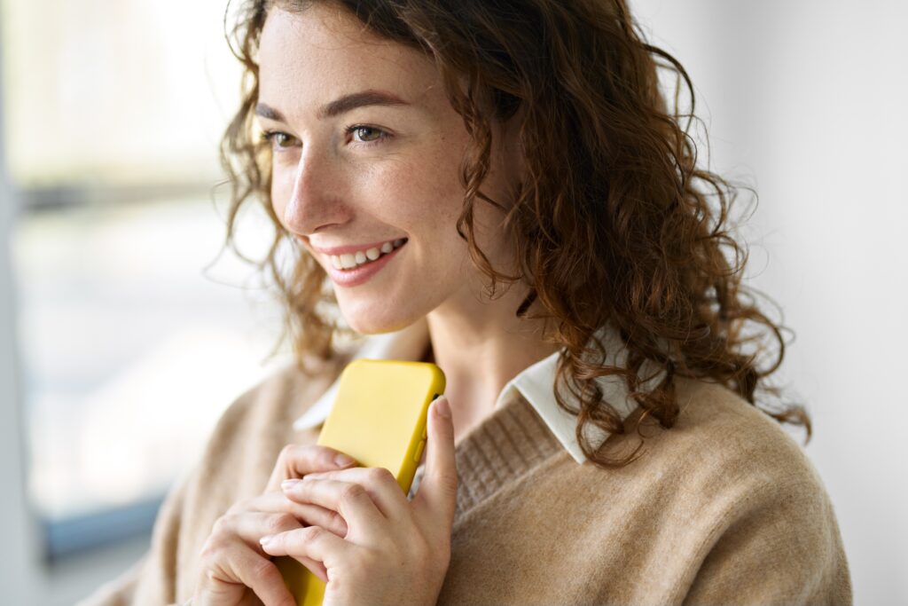 Smiling woman holding a smartphone indoors, representing an engaged consumer participating in online surveys through the LEO platform and its specialized research panels.