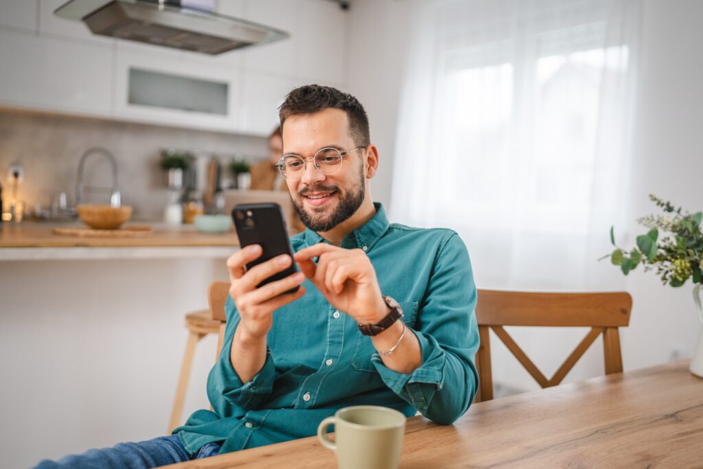 Homme souriant assis à une table de cuisine, utilisant son smartphone pour découvrir la plateforme LEO ainsi que les concours et récompenses en cours.