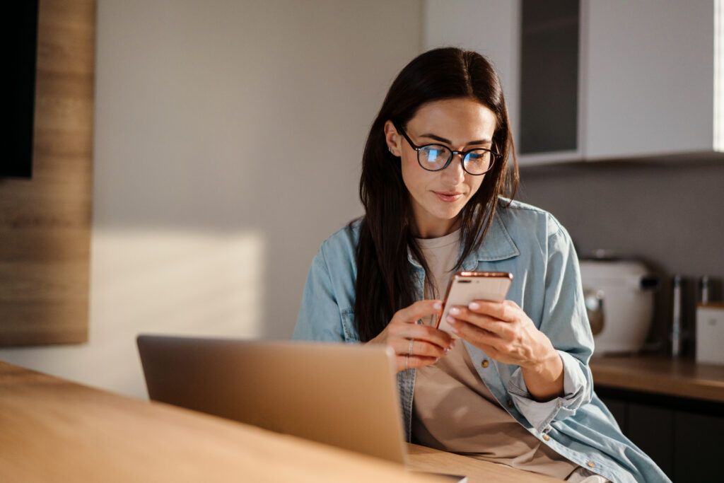 Woman wearing glasses sitting at a kitchen table with a laptop, using her smartphone while participating in LEO contests.