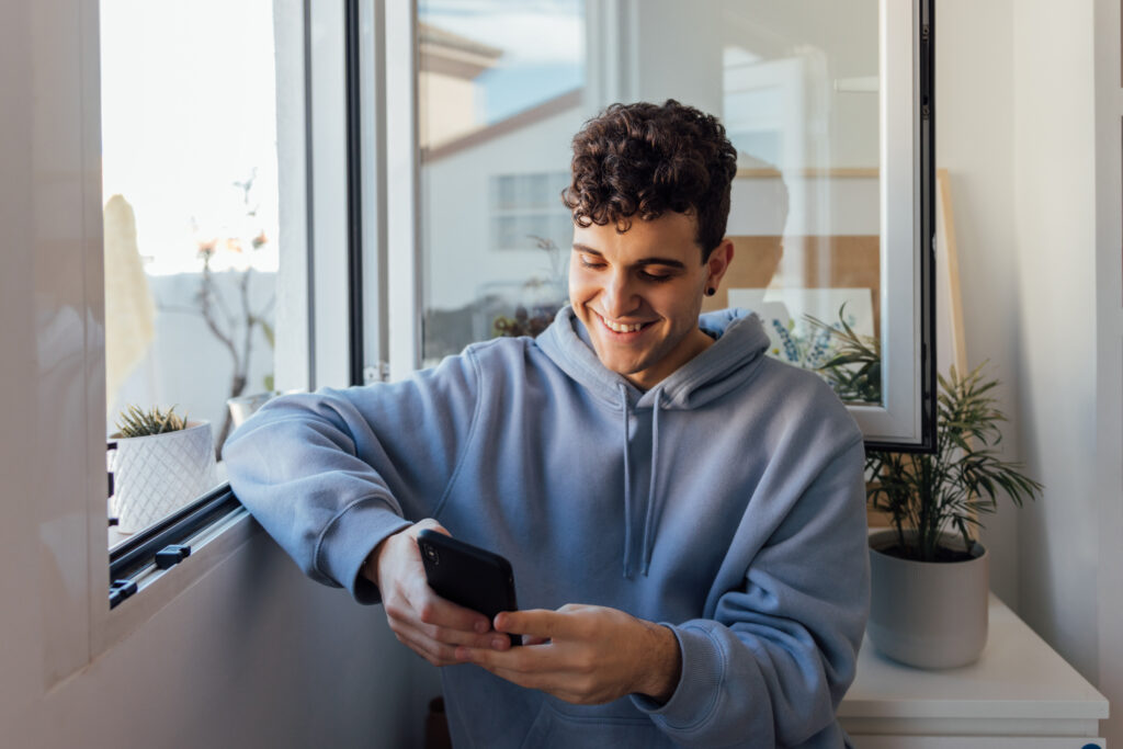 Smiling young man in a blue hoodie sitting by a window at home, using his smartphone while participating in LEO contests.