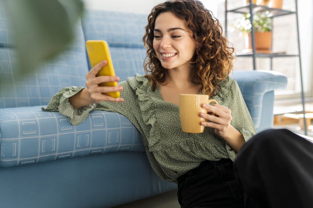 Femme souriante détendue au sol près d’un canapé, tenant un café et utilisant son smartphone tout en explorant la plateforme de médias sociaux de LEO.