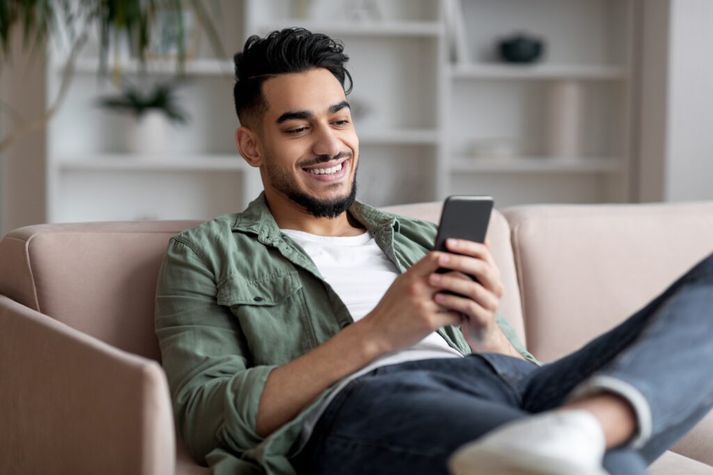 Jeune homme souriant détendu sur un canapé à la maison, utilisant son smartphone tout en explorant la plateforme de médias sociaux de LEO.