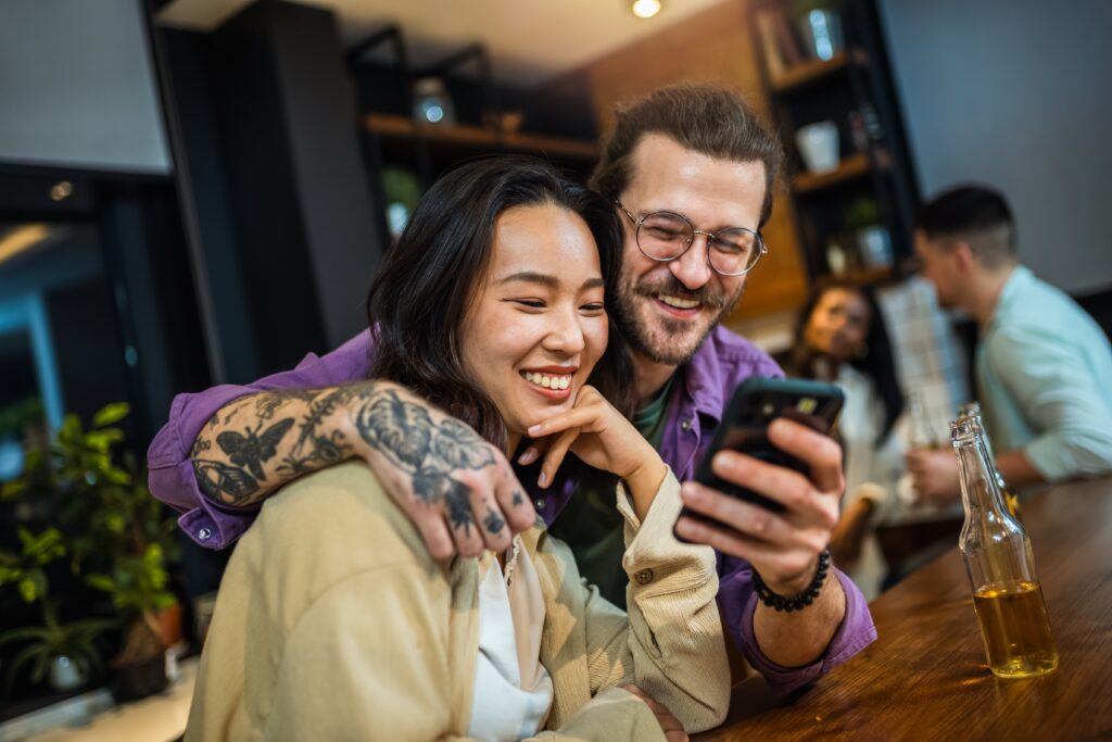 Homme souriant montrant la plateforme LEO sur son smartphone à une amie dans un bar, mettant en avant la promotion Invitez un ami.