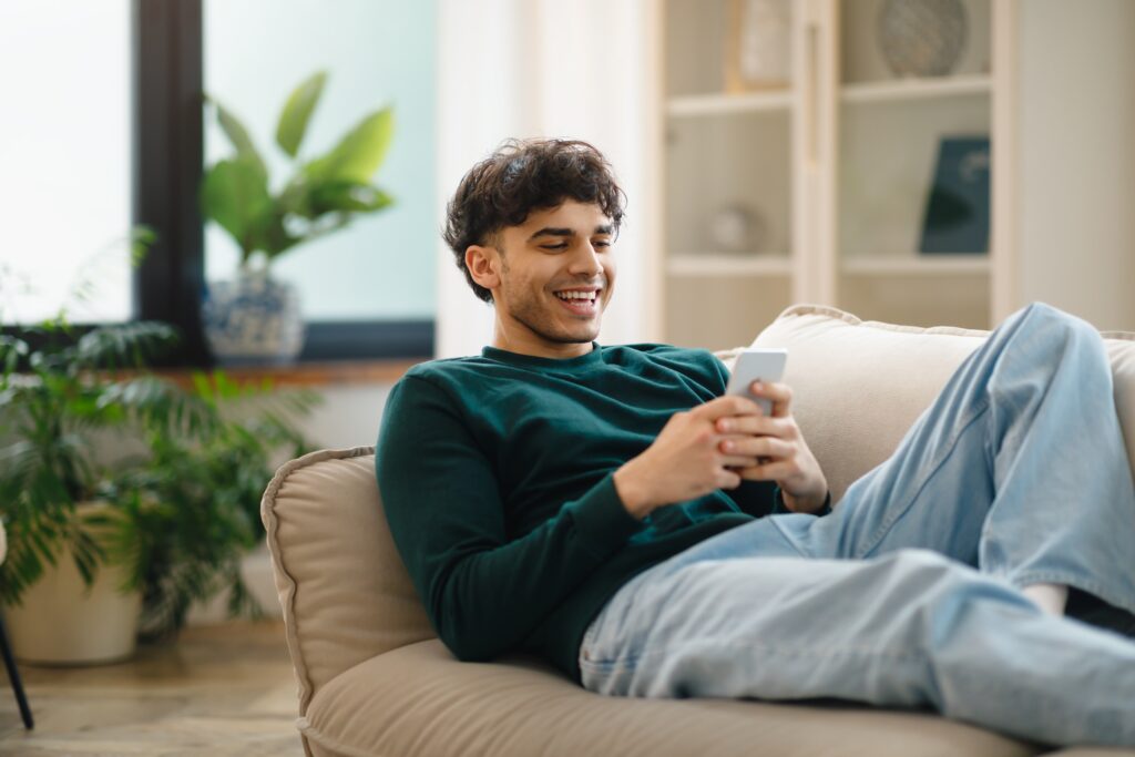 Smiling young man relaxing on a couch at home, using his smartphone while participating in a LEO contest.