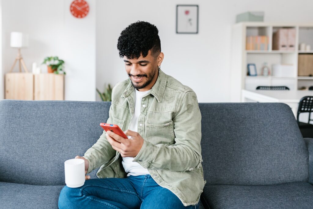 Homme souriant assis sur un canapé à la maison, tenant un café et utilisant son smartphone tout en participant à un concours LEO.