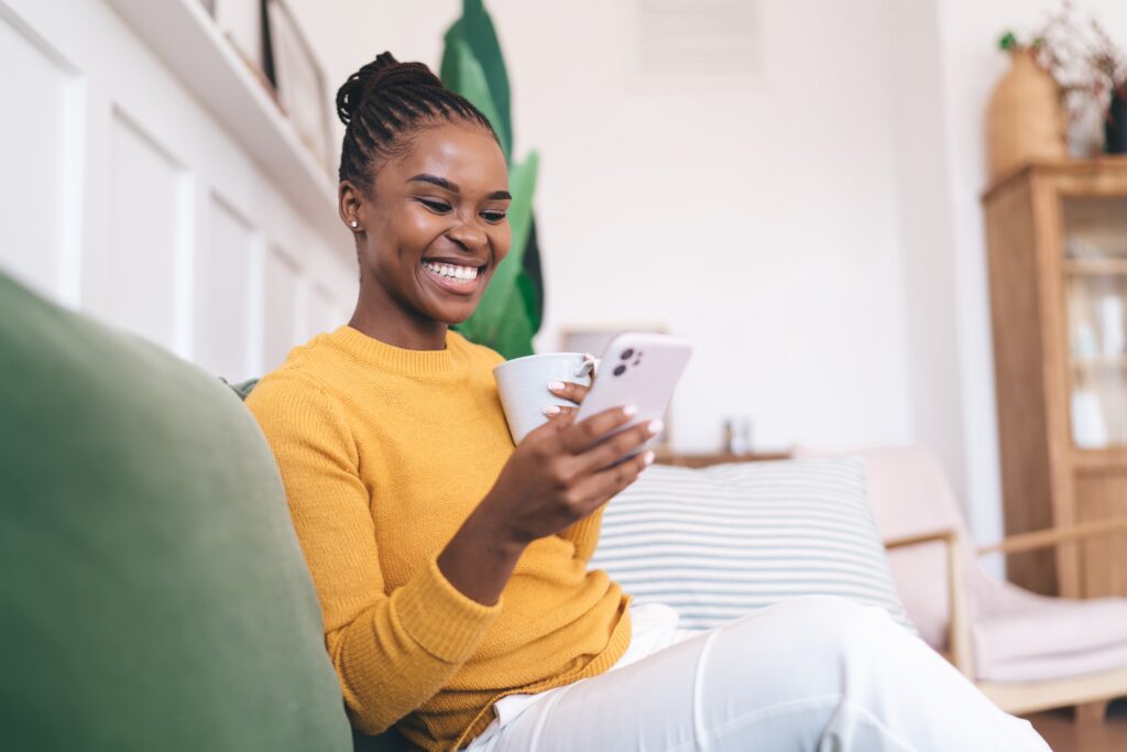 Femme souriante se détendant à la maison avec une tasse de café, utilisant son smartphone tout en participant à un concours LEO.