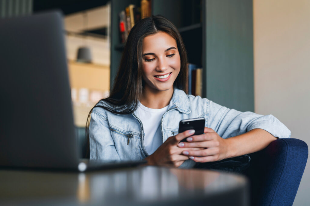 Smiling woman using her smartphone while sitting next to a laptop, participating in a LEO contest online.