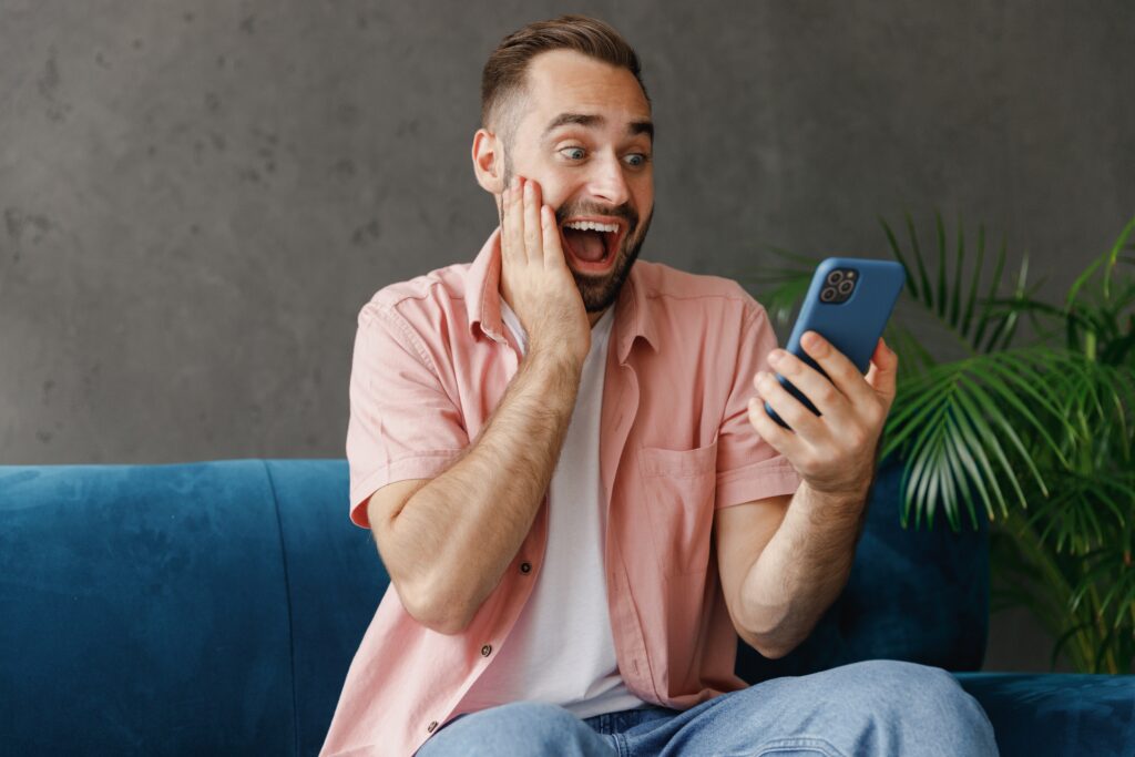 Excited man sitting on a couch, looking at his smartphone and celebrating after winning a prize in a LEO contest.