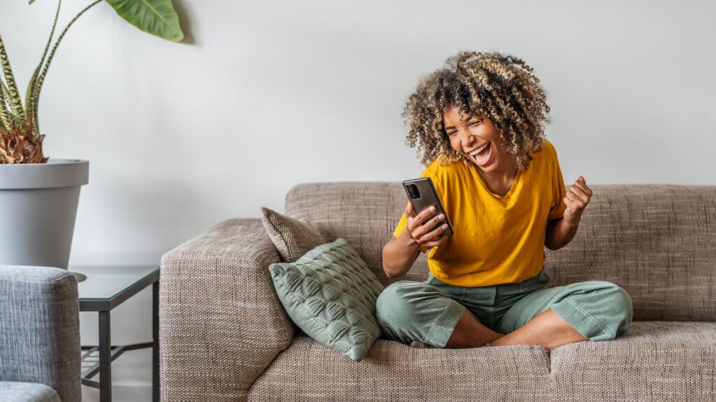 Excited woman sitting on a couch at home, celebrating while looking at her smartphone after winning a prize in a LEO contest.
